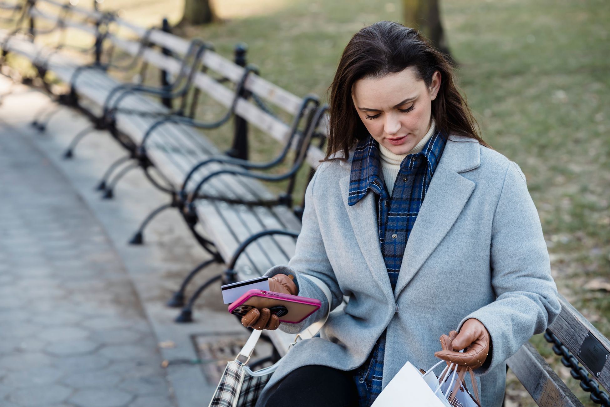 Thoughtful female with brown hair in coat sitting on bench after shopping and holding smartphone and credit card on street in daytime