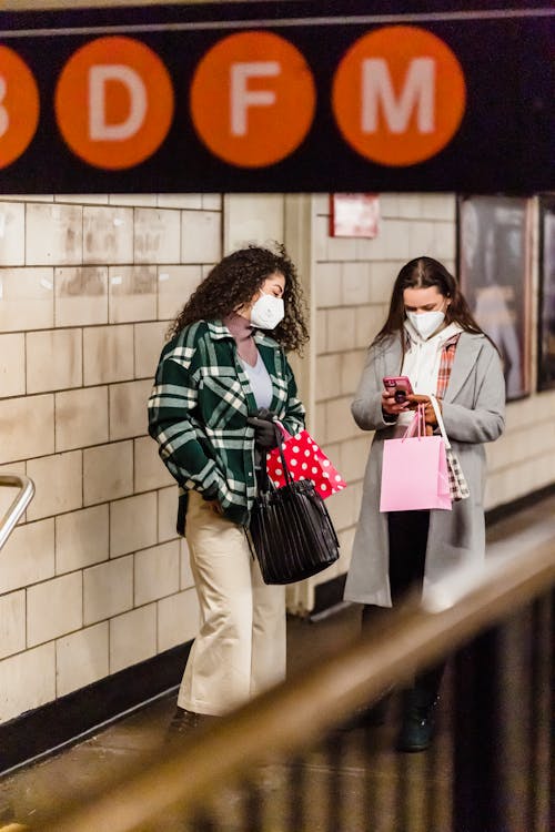 Free Pensive women friends in trendy clothes and sterile masks standing in underground and browsing smartphone during coronavirus Stock Photo