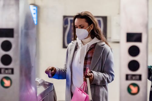 Young woman in warm clothes and protective mask with gloves and shopping bag with purse near turnstile in bright subway station