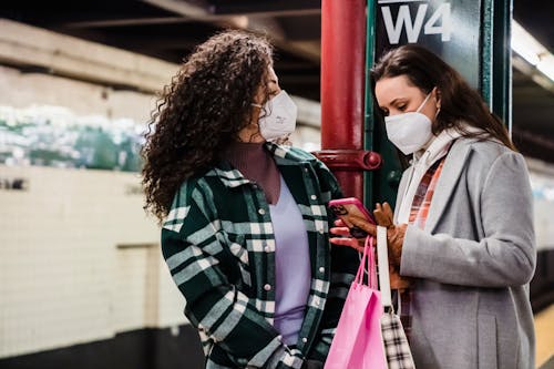 Young women in warm clothes and protective masks standing with shopping bags while browsing on mobile in bright subway station