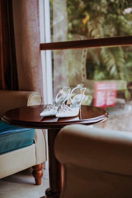 Elegant white bridal shoes placed on round wooden table near comfortable armchairs before wedding ceremony