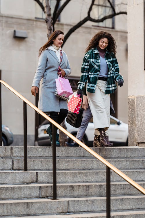 Free Full body of young positive female friends in trendy warm outfit walking downstairs with shopping bags and talking Stock Photo