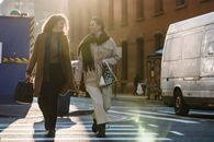 Full length of smiling elegant women in trendy outerwear carrying bags and chatting while crossing carriageway by zebra and looking at each other