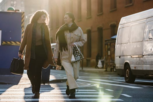 Full length of smiling elegant women in trendy outerwear carrying bags and chatting while crossing carriageway by zebra and looking at each other