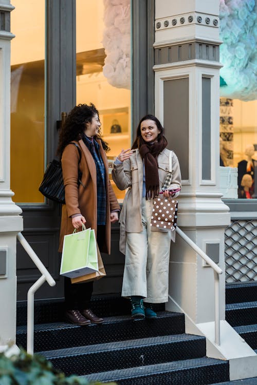 Full body of cheerful female friends laughing and gesticulating while discussing purchases on stairs of store porch