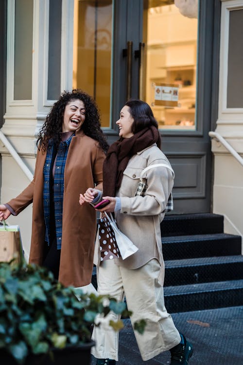 Free Young positive female friends with shopping bags speaking and laughing happily and looking at each other while strolling after visiting shop Stock Photo