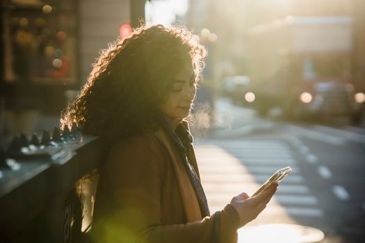 Side view of young female with curly hair touching screen of smartphone near modern cafe on sunny day in back lit