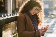 Side view of young female with long curly hair using smartphone intensively on city street on sunny weather