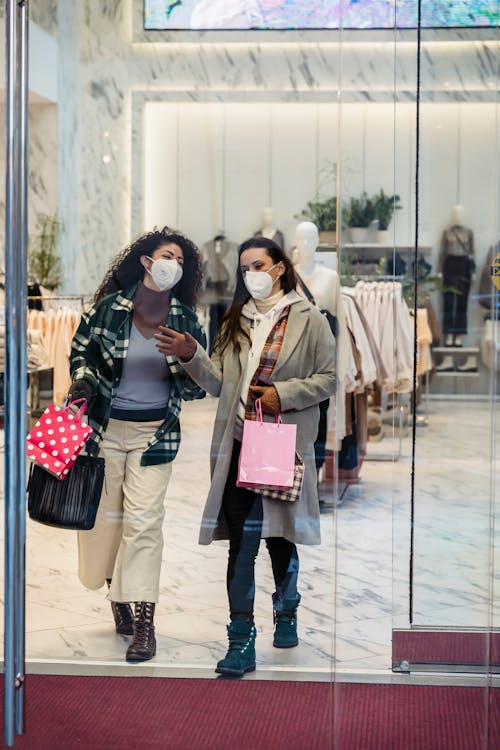 Full body of female friends in protective masks with shopping bags walking through glass doors of clothing store