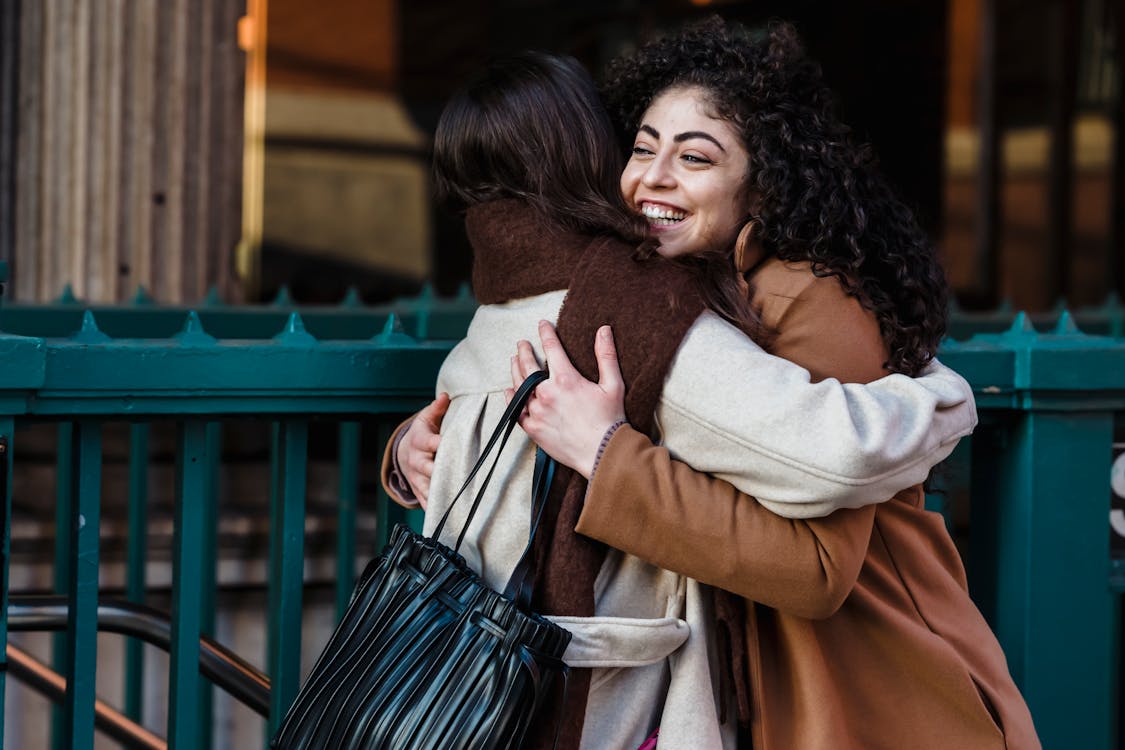 Side view of young happy female friends embracing with pleasure while greeting each other on street