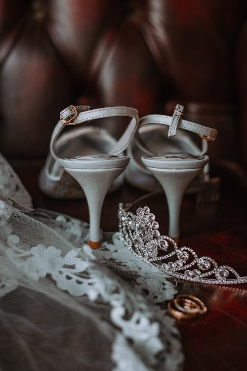 Elegant bridal high heeled shoes and veil with golden rings and tiara placed on leather sofa before wedding celebration