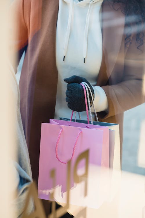 Free Through glass high angle of crop anonymous female buyer with colorful shopping packets standing near showcase of shop Stock Photo
