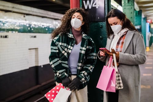 Female in protective mask using mobile phone while waiting for train on platform of subway station with friend standing with gift bags