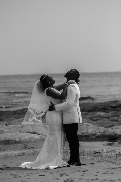 Free Side view of black and white happy young African American bride and groom embracing and smiling while standing on sandy beach near sea on wedding day Stock Photo