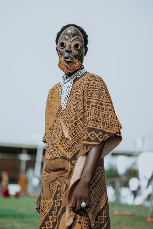 Free Anonymous ethnic male in mask and traditional apparel with ornament and accessories on meadow in town Stock Photo