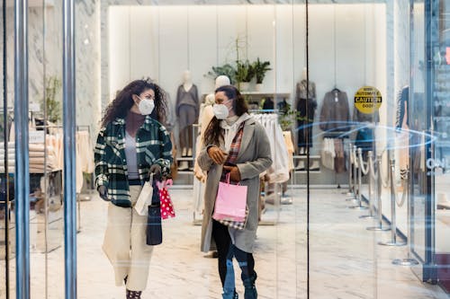 Free Through glass of female friends in protective masks carrying shopping bags while walking out of clothing store Stock Photo