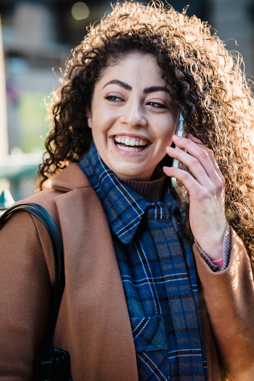 Optimistic young female with curly hair smiling talking on mobile phone and laughing happily