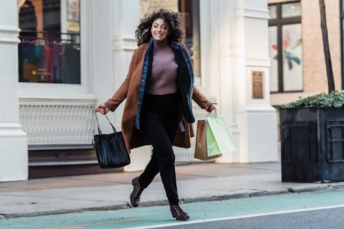 Full body of smiling ethnic female in stylish outfit with shopper bags in hands running on road near building in city