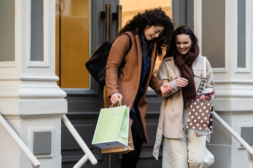 Positive multiracial female friends in elegant wear with gift bags in hands walking out of shop on street in city