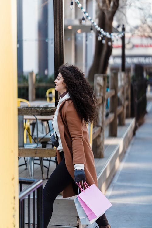 Side view of positive ethnic female with black hair in trendy outfit strolling on street with gift bags in city