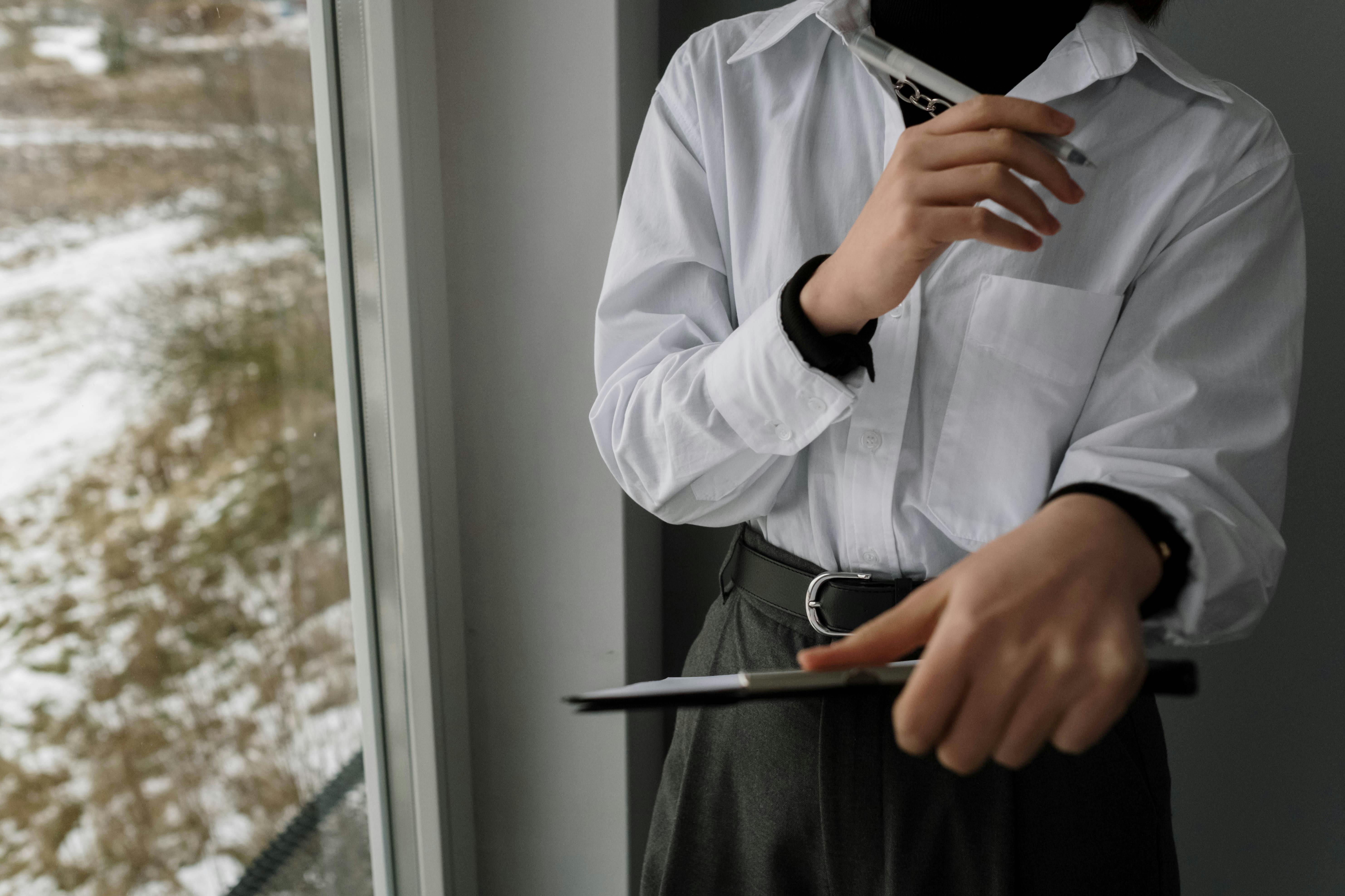 Person in white shirt holds a clipboard near a window, suggesting a professional setting.