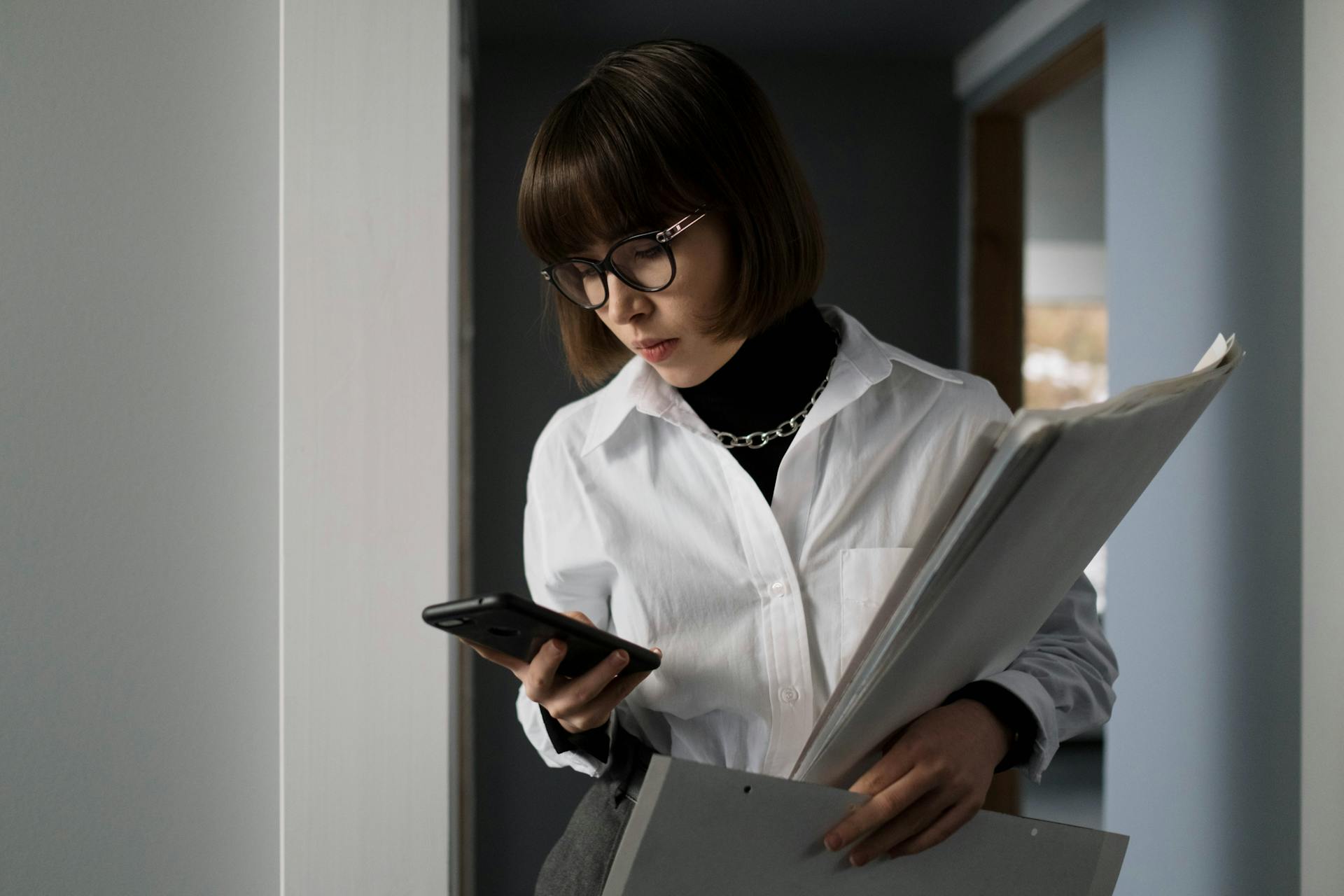 Young woman with eyeglasses, holding documents, checking smartphone indoors.