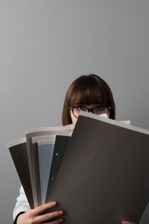 Free A young woman wearing glasses holds large sample papers, partially obscuring her face. Stock Photo