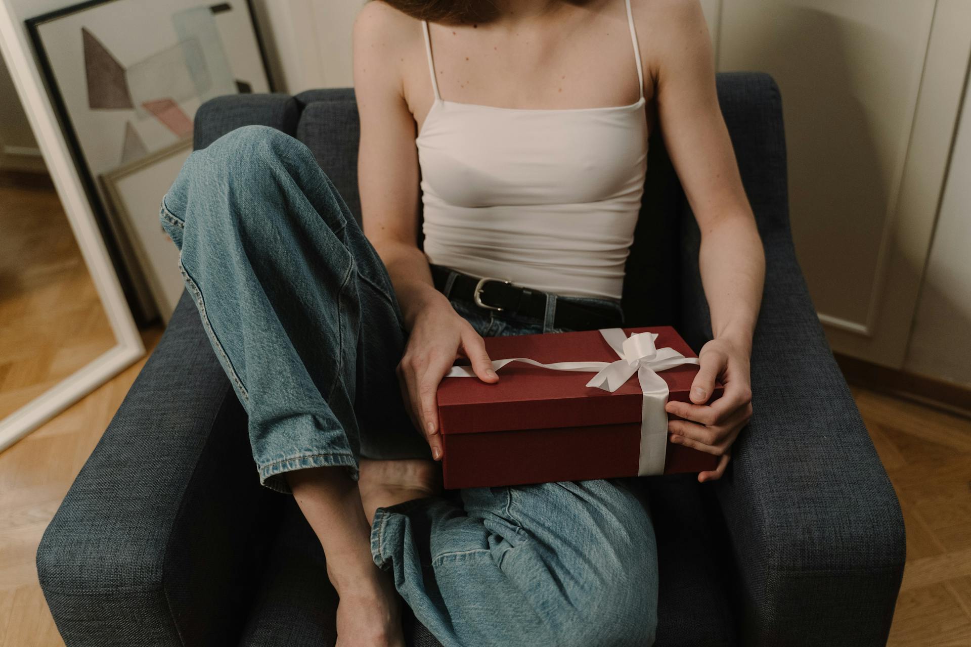 Young woman in casual attire holding a gift box with white ribbon while sitting in a modern armchair.