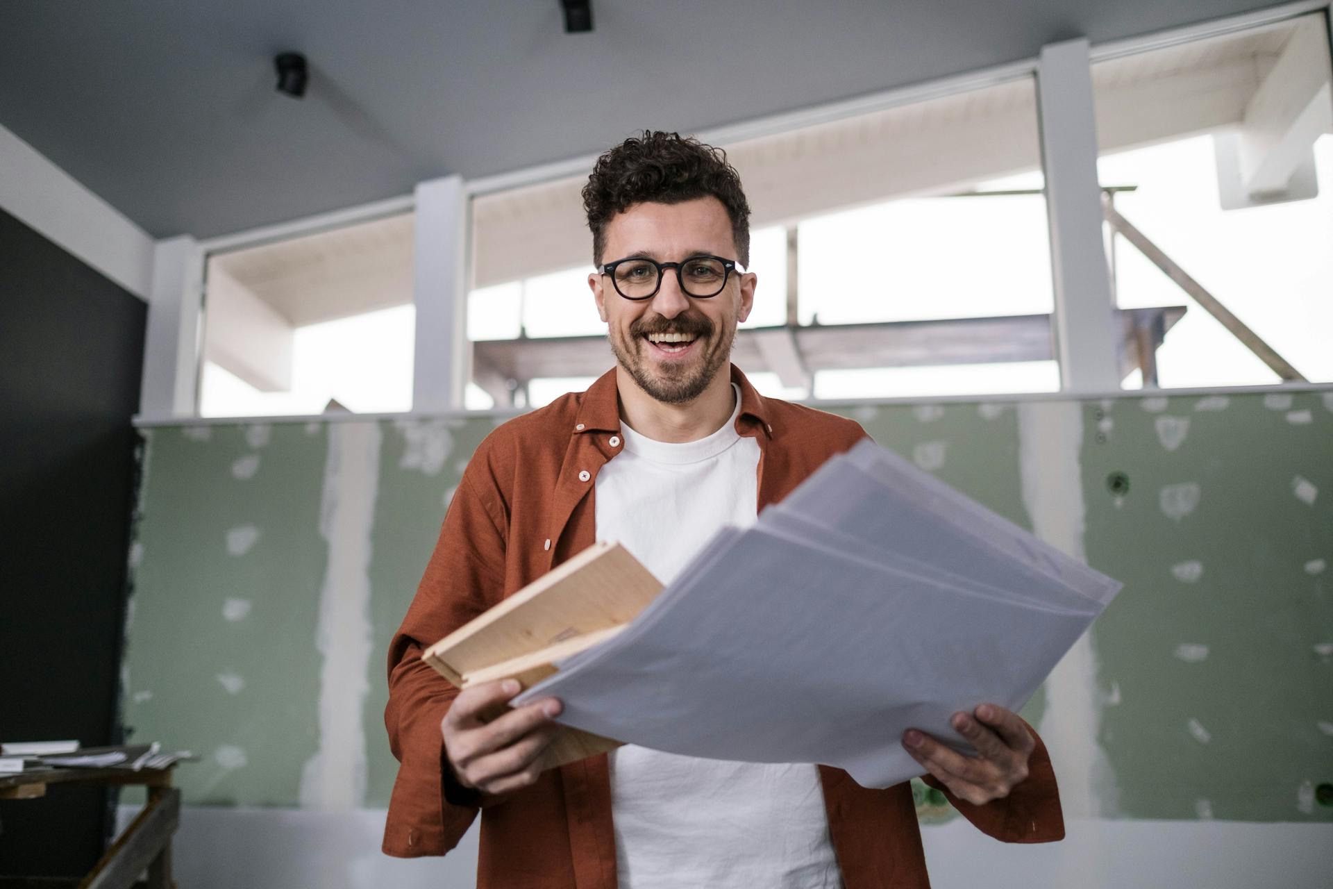 Smiling interior designer holding plans in a partially renovated room, conveying excitement and creativity.