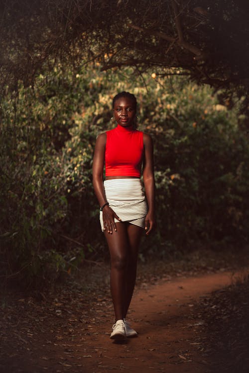 Full body of young African American female millennial in trendy top and mini skirt walking on narrow pathway among lush green trees in forest
