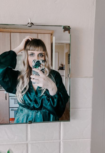 Young calm female in elegant dress touching long blond hair while taking mirror selfie on mobile phone at home