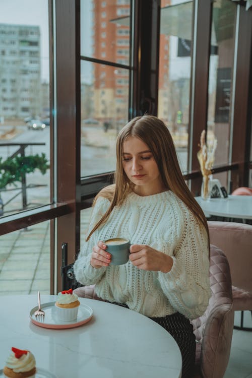 Free Pensive female in sweater drinking hot latte while sitting near window at table with cupcake in modern cafe during coffee break Stock Photo