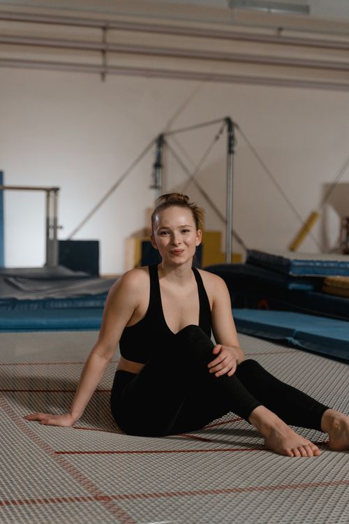 Free A young woman in activewear sitting barefoot on a mat in a gymnastics studio, smiling warmly. Stock Photo
