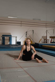 Caucasian woman sitting barefoot on a trampoline in a gym, wearing activewear and smiling.