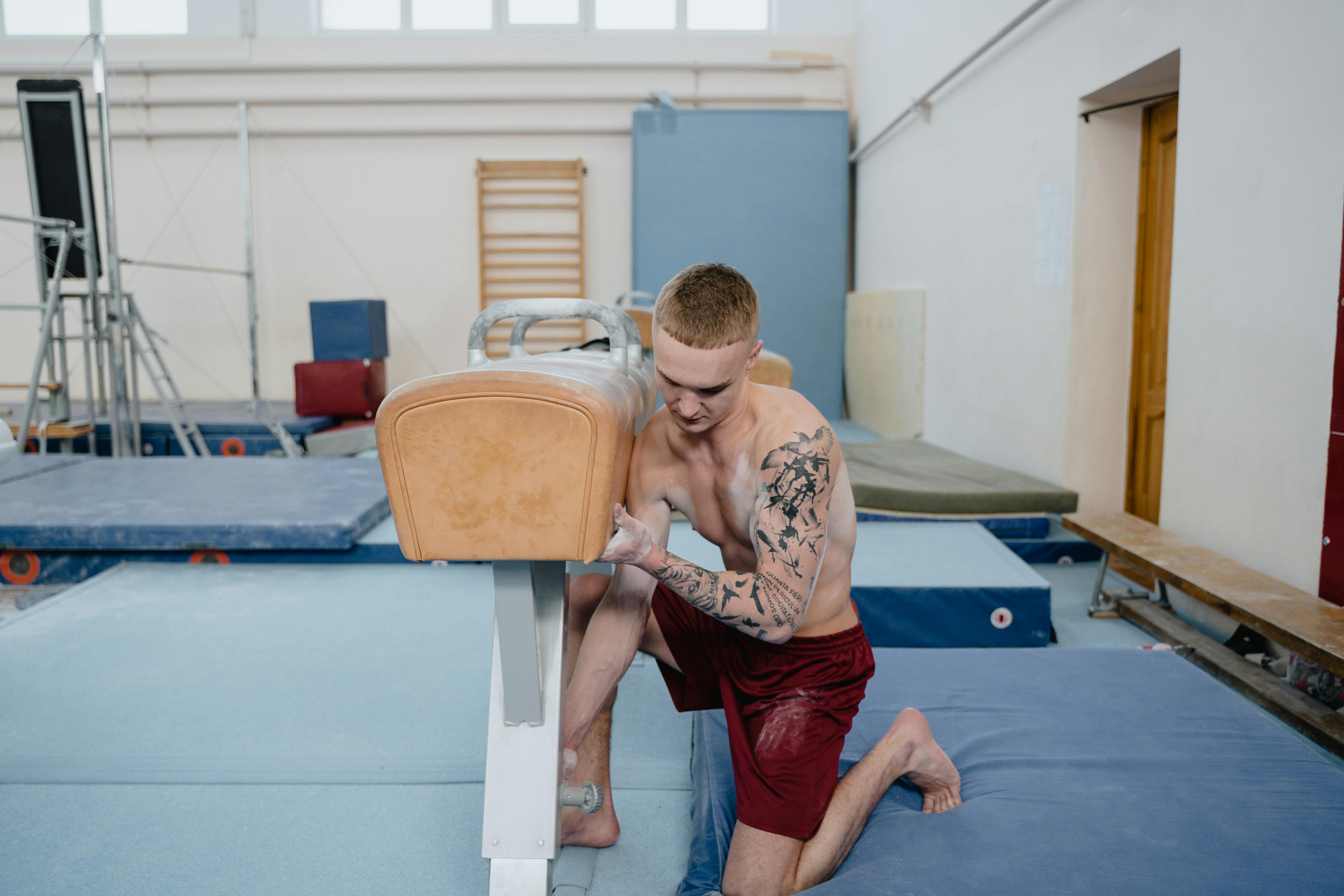 Shirtless male gymnast setting up a pommel horse for training in an indoor gym.