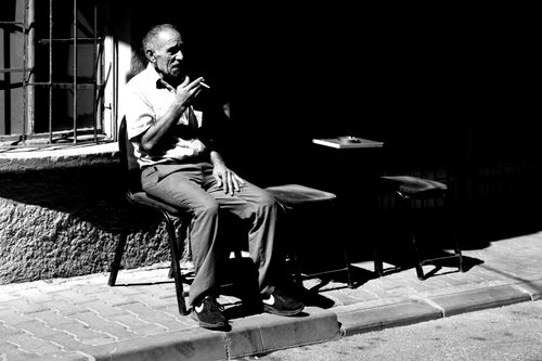 Free Senior man smoking while sitting outdoors in a monochrome street scene. Stock Photo