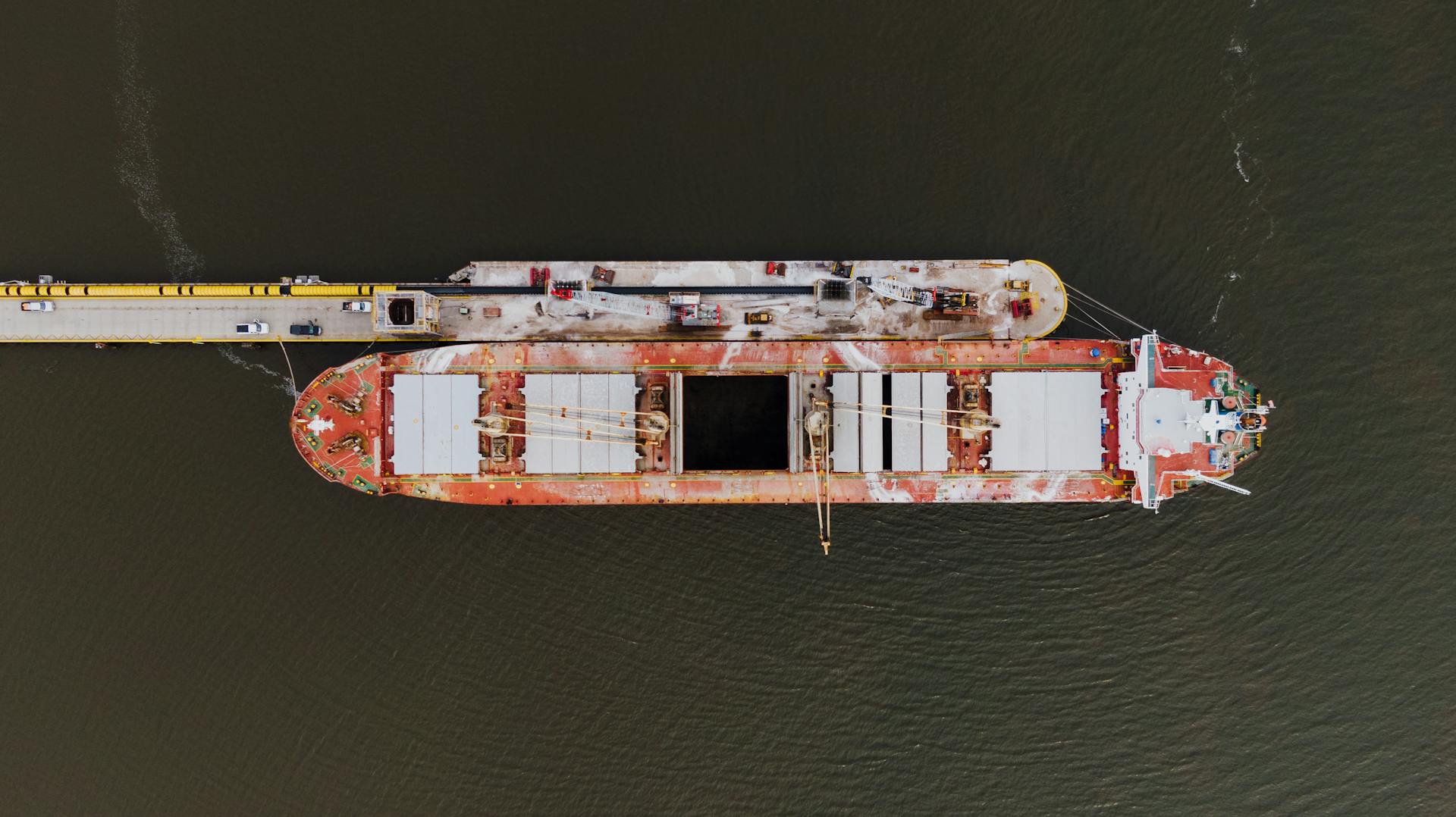 Drone view of barge moored on rippled ocean near dock with vehicles in harbor