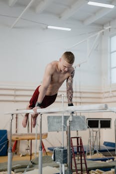 Shirtless male gymnast practicing on parallel bars in a gym, showcasing strength and focus.