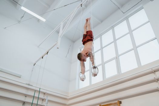 Shirtless gymnast practicing upside down on rings in an indoor gym, showcasing strength and focus.