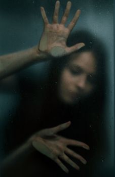 Blurred woman pressing hands against wet glass, creating a mysterious mood.