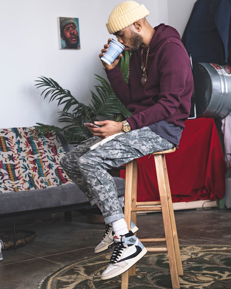 Young Fashionable Man Sitting On A Stool, Drinking Coffee And Scrolling Through His Phone 