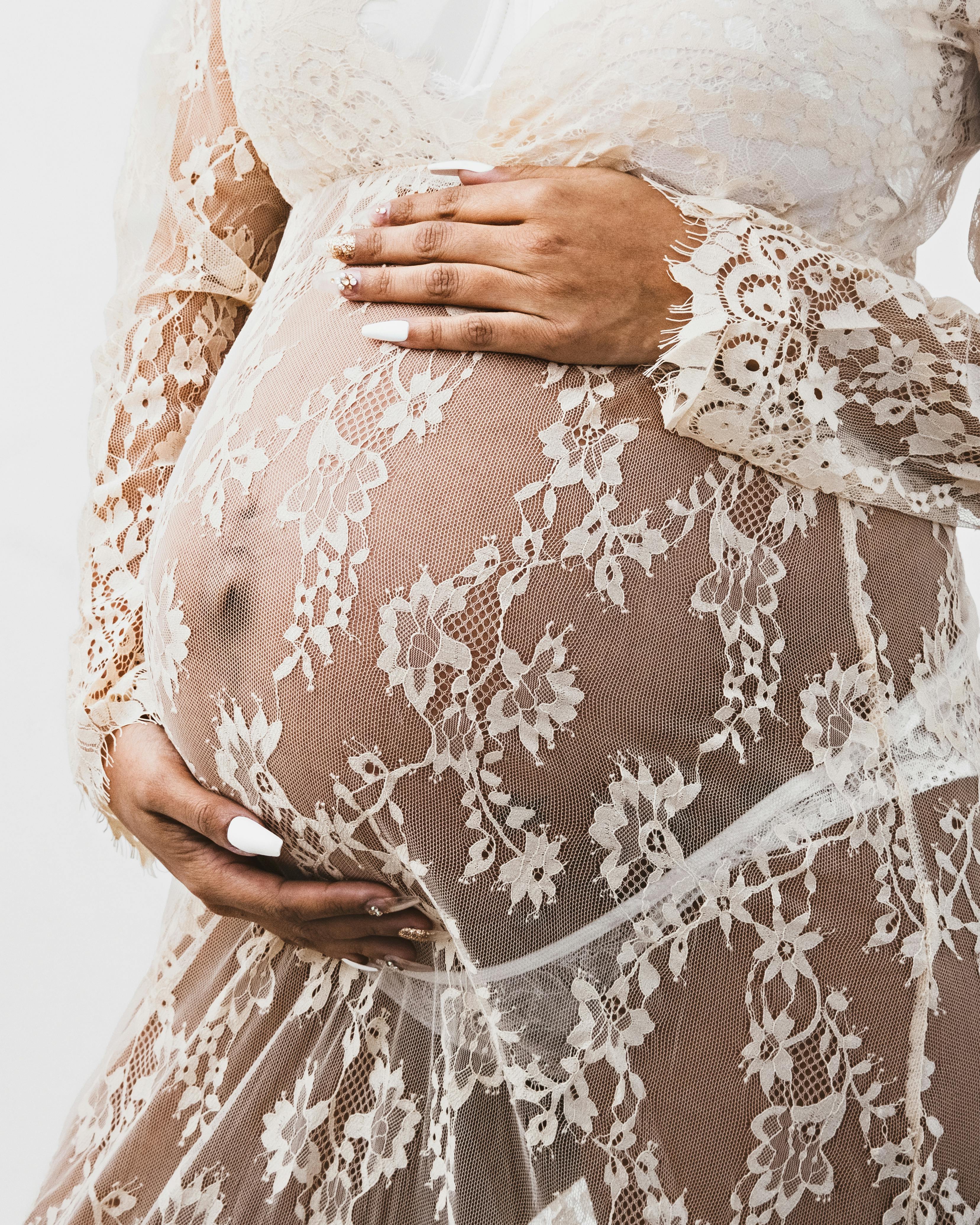 Free Pregnant woman in a sheer lace dress embracing her belly with elegance. Stock Photo