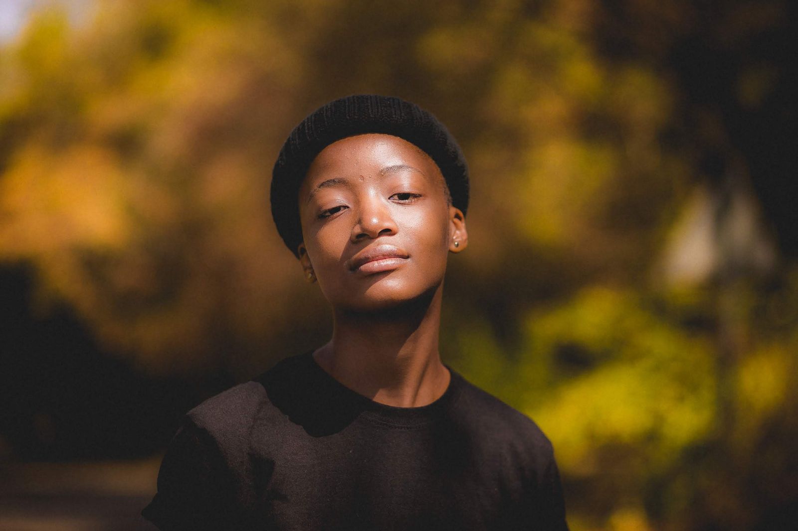 A Black teenager in a black beanie and T-shirt looks toward the camera in warm sunlight, with blurred autumn trees behind.