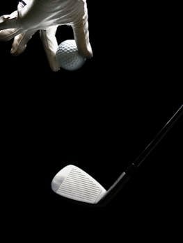 Golf ball held by a gloved hand with a golf iron against a dark background.