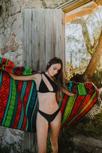 Young woman poses outdoors in a black bikini, draped with a vibrant, colorful blanket for a stylish summer fashion look.