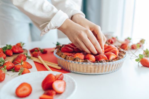 Hands skillfully arrange fresh strawberries on a homemade tart in a bright kitchen setting.