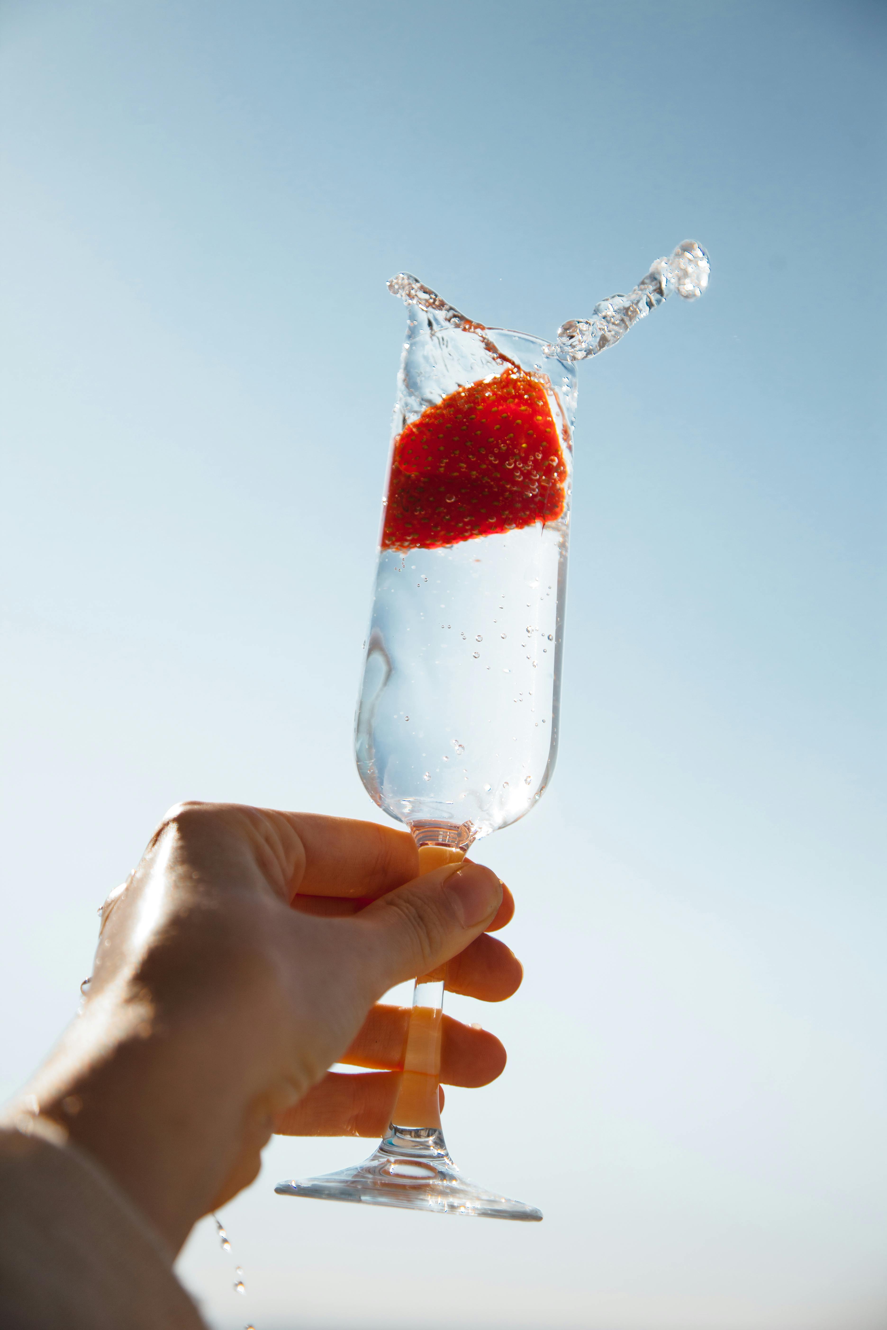A hand holding a glass with a strawberry and sparkling water splash against a clear sky.