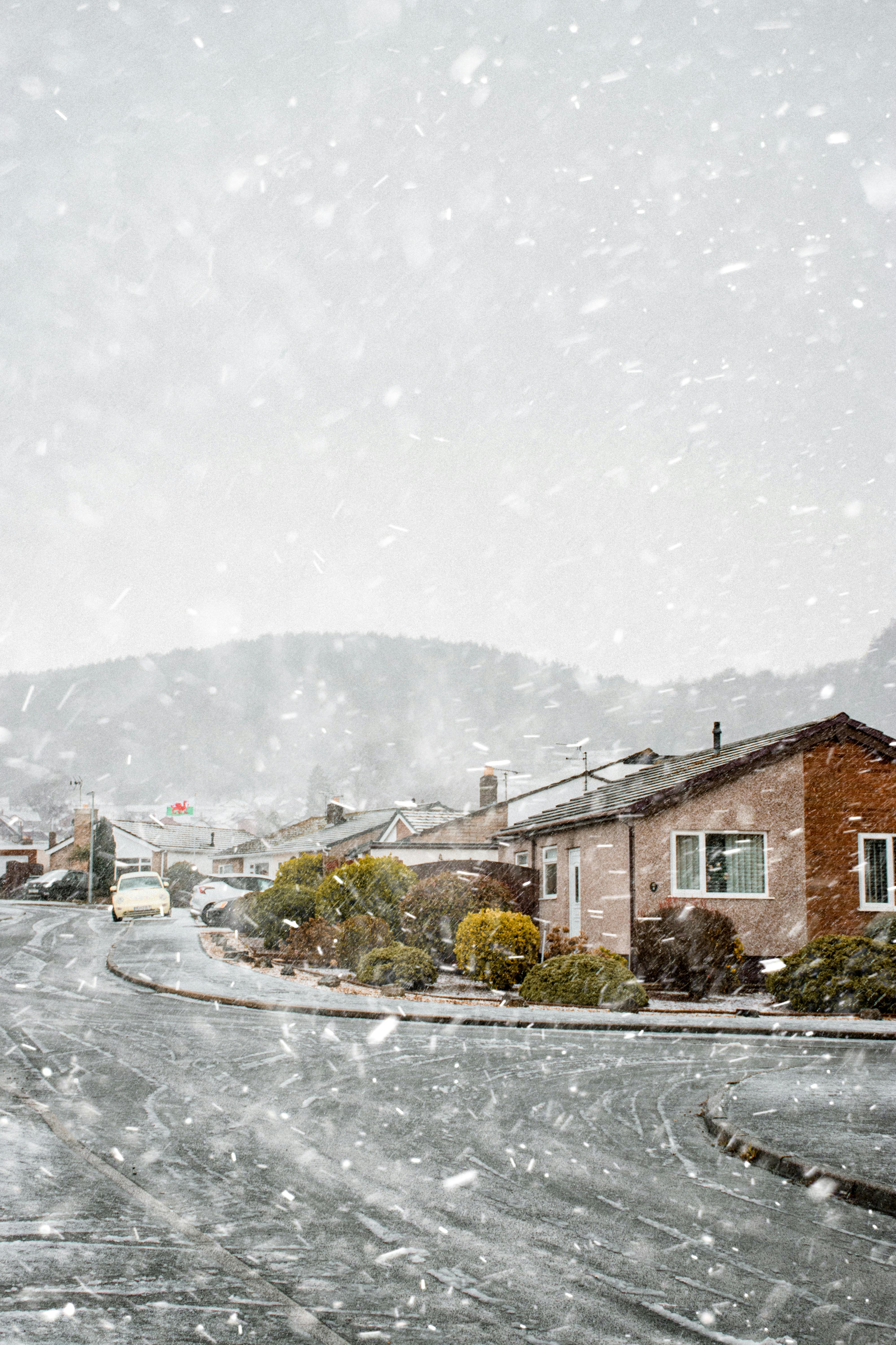 A serene winter scene of houses blanketed in snow with snowflakes gently falling on a quiet street.