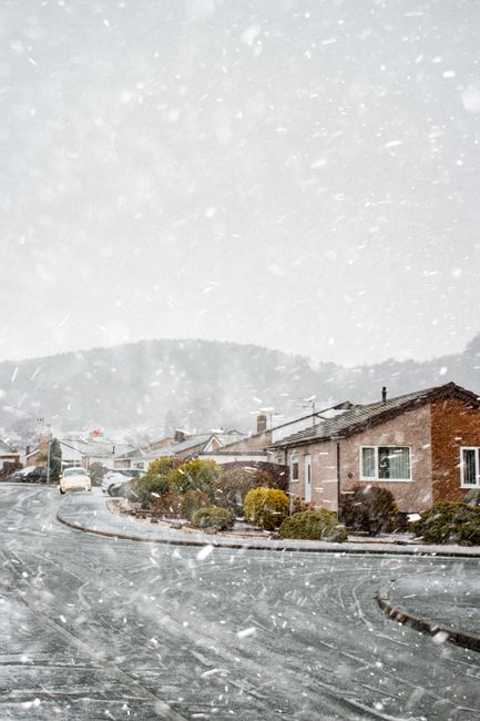 A serene winter scene of houses blanketed in snow with snowflakes gently falling on a quiet street.