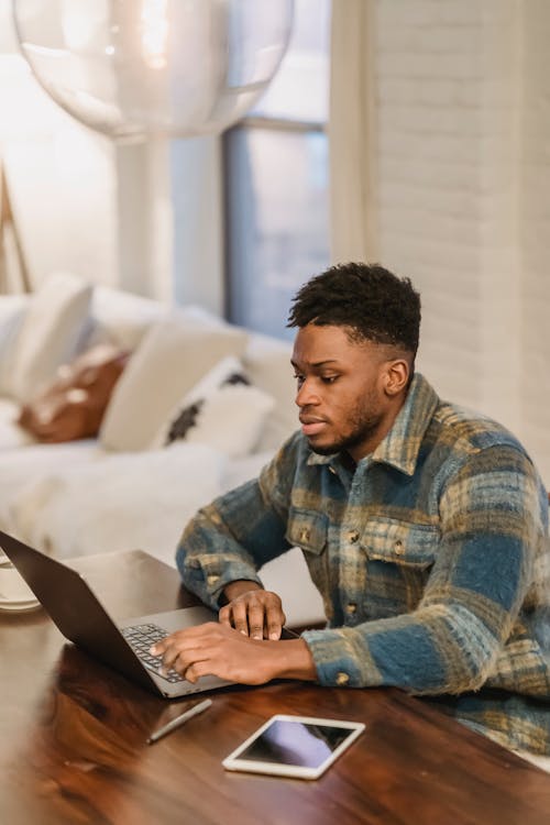 Free Focused bearded African American male freelancer in casual clothes sitting at table with laptop tablet and coffee while working on remote project at home Stock Photo